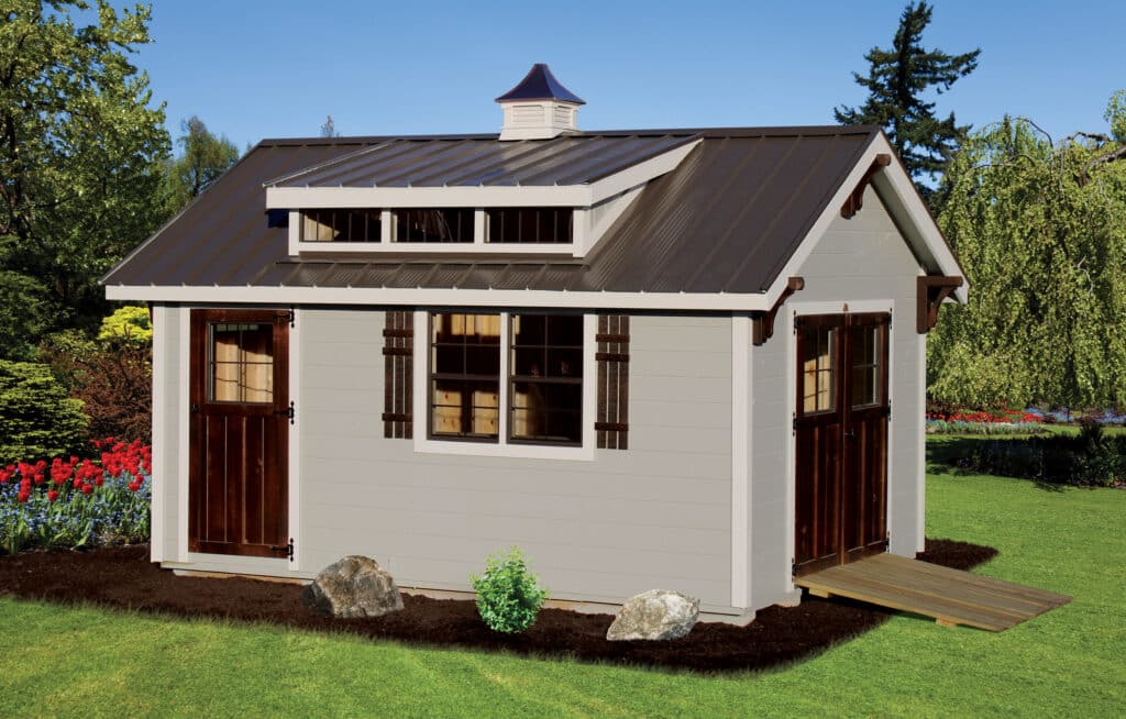 gray utility shed with dormer and cupola on brown metal roof and two brown doors with one on the side with ramp leading up to it
