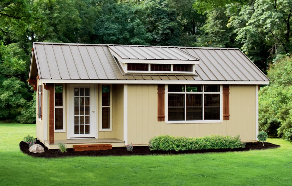tan gable shed with front porch dormer on metal roof and white trim surrounded by mulch garden bed