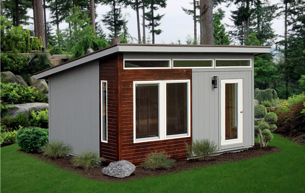 gray studio shed with brown wood detailing featuring large windows and slated roof and garden bed around its perimeter