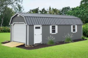 Gray lofted garage shed with white garage door, walk-in door, trim, and shutters. Wooden ramp sits between garage door and grass