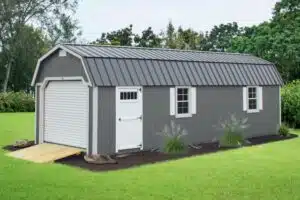 Gray lofted garage shed with white garage door, walk-in door, trim, and shutters. Wooden ramp sits between garage door and grass